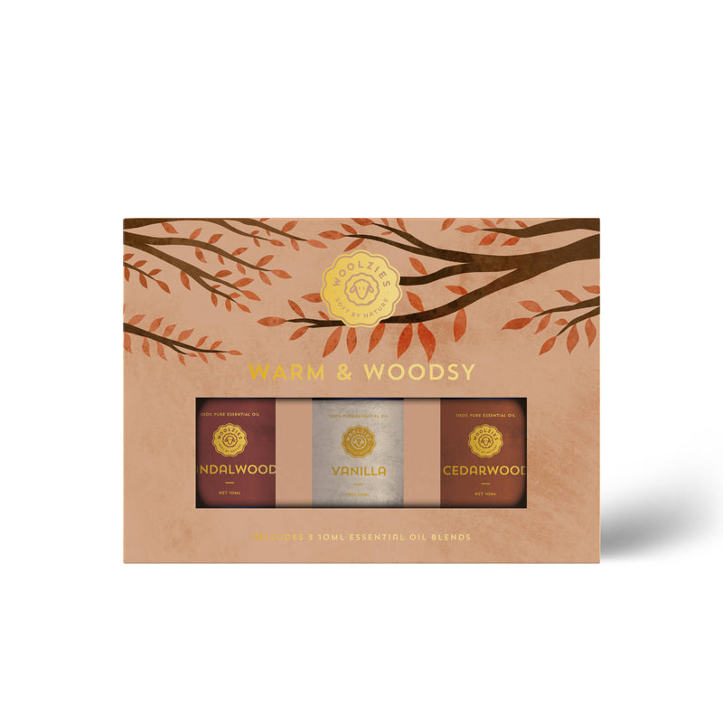 The Woolzies Warm & Woodsy Collection features three 10ml bottles—sandalwood, vanilla, and cedarwood essential oils—packaged in a box with brown and orange leafy designs and a gold circular Woolzies logo on top.