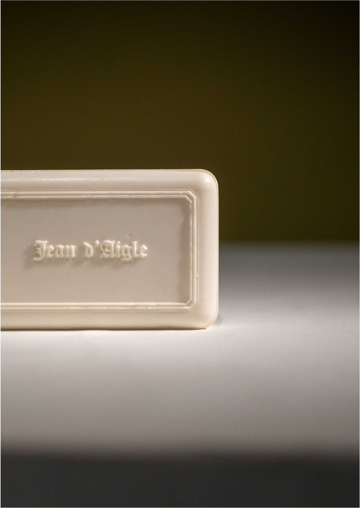 Close-up of a white soap bar with the text "Jean d’Aigle" engraved on it. The handcrafted Jean d'Aigle Jasmine Soap, infused with a delicate jasmine scent, has smooth edges and rests on a neutral-colored surface with a dark, out-of-focus background. Only the right portion of the soap is visible in the image.