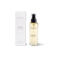 A clear bottle of Archipelago Botanicals Bergamot Eucalyptus Hydrating Body Oil with a black pump sits next to its white box, both featuring the product name and details in elegant gold lettering.