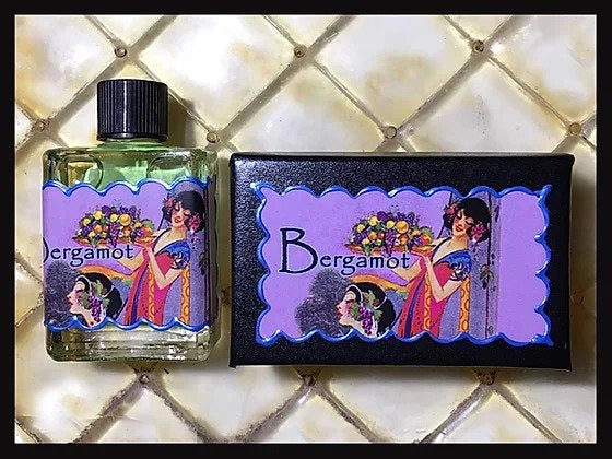 The "Seventh Muse Fragrant Oil - Bergamot" by Seventh Muse Fragrant Oils features a bottle and matching box with art of a woman in colorful attire holding flowers. The cruelty-free perfume oil's design is set against a quilted, cream-colored background.