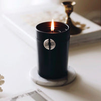 The Archipelago Black Forest Boxed Candle by Archipelago Botanicals glows on a white marble coaster, beside a gold candlestick and an open book or magazine, all set atop a light surface.