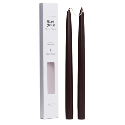 Two tall, dark brown Archipelago Black Forest Tapered Candles by Archipelago Botanicals stand upright next to a white rectangular box labeled “Black Forest Special Reserve,” which features a window displaying the scented candles inside.