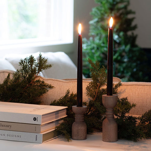 Two lit Archipelago Botanicals Archipelago Black Forest Tapered Candles in wooden holders sit on a table with evergreen branches beside books, in a cozy room with a sofa and a decorated Christmas tree in the background.