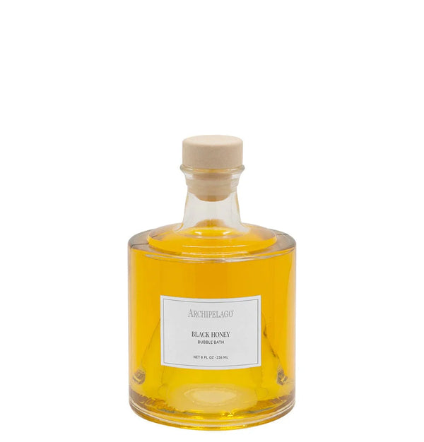 A clear glass bottle filled with yellow liquid, sealed with a beige cap, and labeled "Archipelago Botanicals Black Honey Bubble Bath" offers skin-softening ingredients and a luxurious bath experience from Archipelago Botanicals.