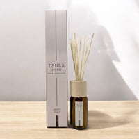 A bottle of ISULA parfums Room Diffuser - Dolce Farniente No.28 with wooden cap and reeds stands beside its matching box, bringing a Mediterranean scent to light surroundings, set against a softly lit background.