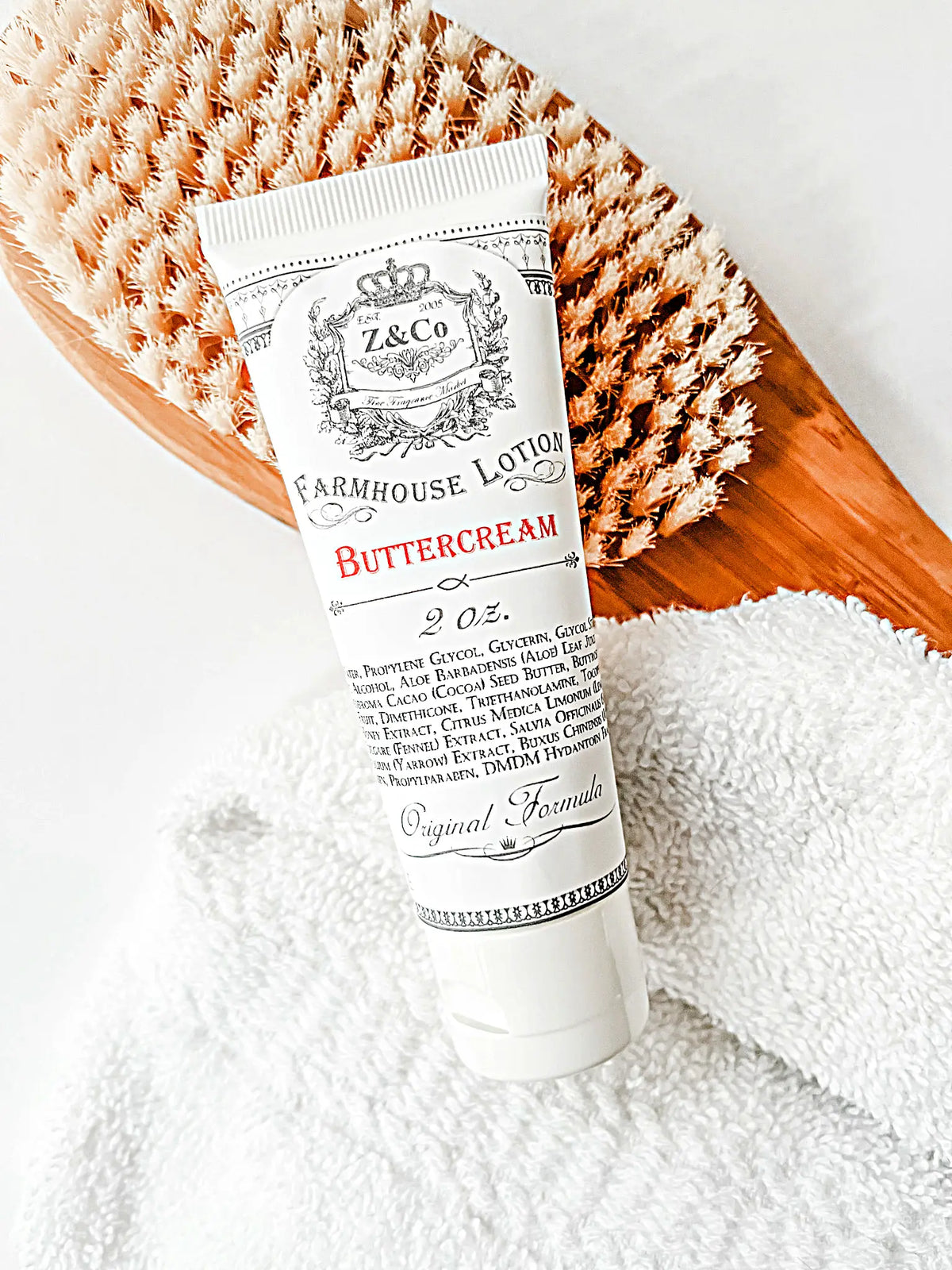 A tube of Z&Co. Buttercream Farmhouse Lotion sits on a white towel next to a wooden bath brush with bristles. This travel-sized lotion, featuring the original buttercream formula, comes in a 2-ounce size, and the image exudes a bright, clean aesthetic.