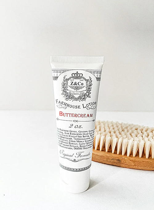 A 2 oz. tube of Z&Co. Buttercream Farmhouse Lotion stands upright, showcasing elegant black text and intricate design elements on its white surface. Highlighting its buttercream formula, a wooden brush with white bristles lies horizontally in the background.