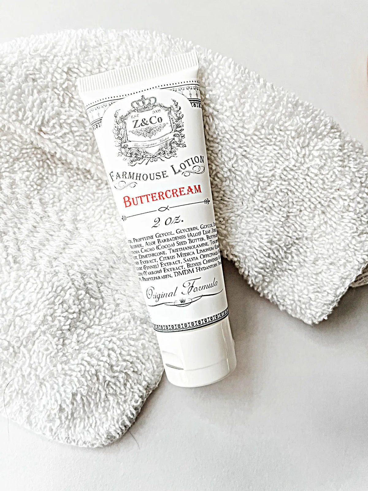 A tube of Z&Co. Buttercream Farmhouse Lotion Tube (2 oz.) resting on a soft white towel. The lotion container is adorned in white with elegant, vintage-style black and red text detailing ingredients and product information. Enjoy the luxurious buttercream formula in this travel-friendly size wherever your journeys take you.