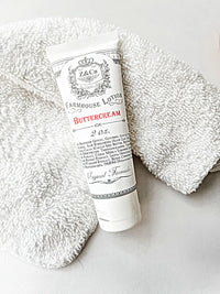 A tube of Z&Co. Buttercream Farmhouse Lotion Tube (2 oz.) resting on a soft white towel. The lotion container is adorned in white with elegant, vintage-style black and red text detailing ingredients and product information. Enjoy the luxurious buttercream formula in this travel-friendly size wherever your journeys take you.