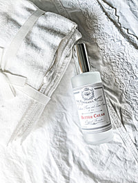 A bottle of the "Z&Co. Buttercream Farmhouse Fall Room/Linen Spray" from Z&Co. rests on a textured white cloth surface. Beside it lies a neatly folded white cloth tied with a white ribbon, boasting intricate detailing. This long-lasting scent delivers a luxurious aroma perfect for giving your home that cozy farmhouse fall feel.