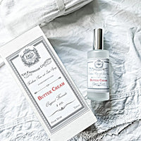 A bottle of "Z&Co. Buttercream Farmhouse Fall Room/Linen Spray" is placed next to its matching box on a textured white fabric background. The label on both the bottle and box displays detailed, vintage-style design with the brand "Z&Co." and other descriptive text, promising a long-lasting scent.