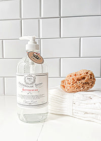 A clear bottle of Z&Co. Buttercream Farmhouse Liquid Soap labeled "Buttercream" with a pump dispenser sits on a white tiled surface. Beside it are a natural sponge and folded white towels, creating a clean and elegant bathroom setup with a delicate scent.