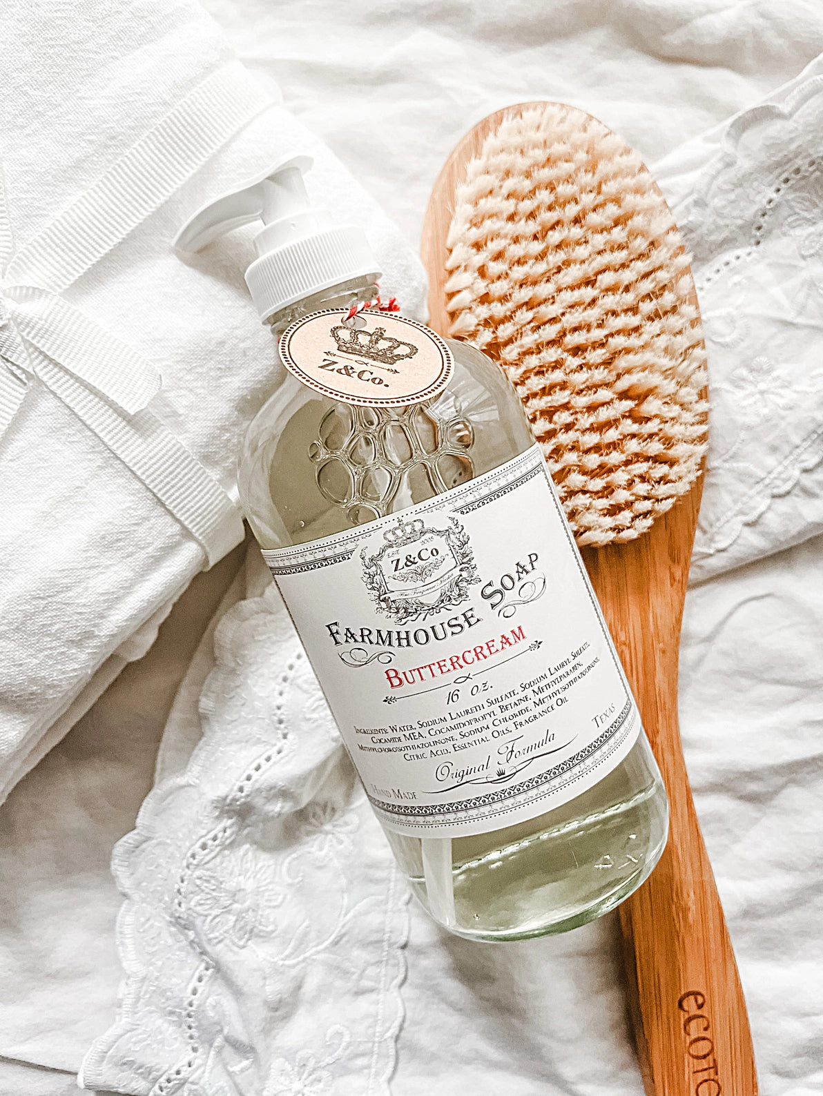 A bottle of Z&Co. Buttercream Farmhouse Liquid Soap lies on white fabric, showcasing its label "Buttercream." Beside it is a large wooden bath brush with light bristles. The scene exudes a clean, minimal, and rustic aesthetic, with small delicate fabrics partially visible in the background, hinting at its delicate scent.