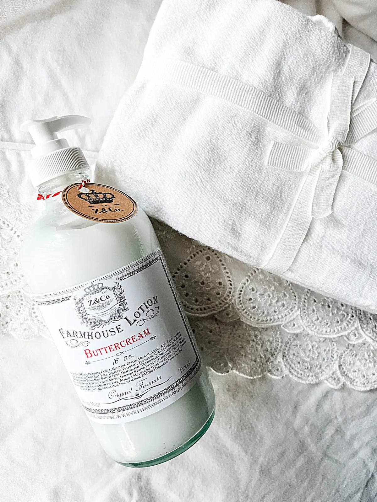 A pump bottle of Z&Co. Buttercream Farmhouse Lotion, celebrated for its non-greasy formula, rests on a folded white lace fabric tied with a ribbon. The bottle features a beige tag attached by a red string, and both items are elegantly placed on a white surface.