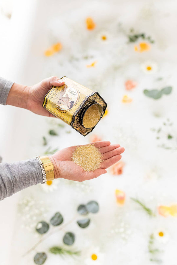 A person pours Barefoot Venus Mustard Bath Soak - 100% Natural into their palm over a floral-decorated tub with milky water, orange and yellow flowers, and green leaves. They wear a gold bracelet.