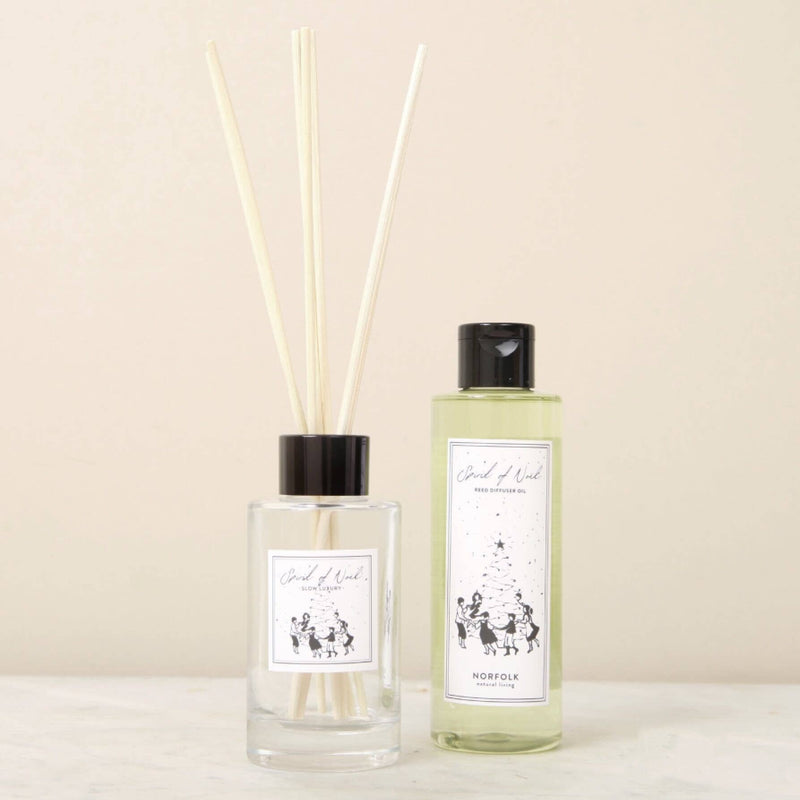 A clear reed diffuser with light wooden sticks and a matching refill bottle labeled "Spirit Of Noel" by Norfolk Natural Living, both filled with essential oils, are displayed on a light surface against a beige background.