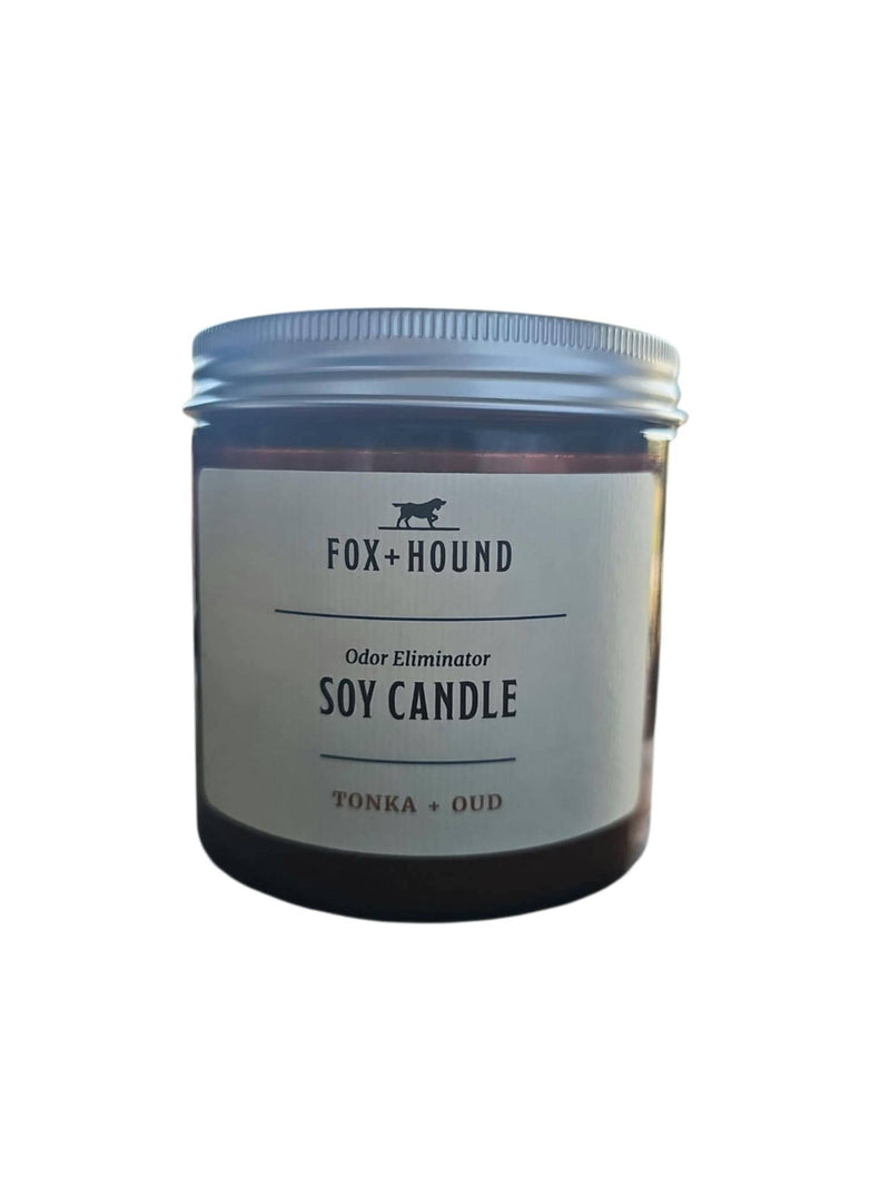 A brown jar with a silver lid, labeled "Fox + Hound Odor Eliminator Soy Candle - Tonka & Oud," effectively neutralizes pet odors; shown against a white background.