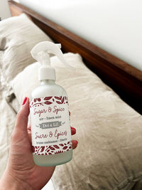 A hand holds a bottle of Dot & Lil Sugar & Spices Linen + Air Mist in front of beige pillows on a neatly made bed with a wooden headboard.