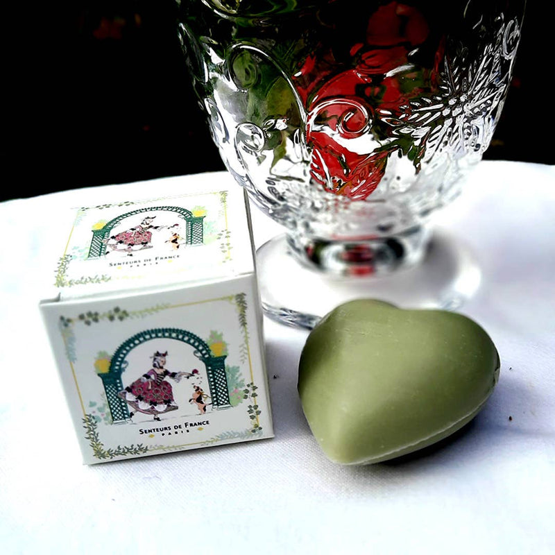 A Senteurs De France Olive Heart Soap - Marquise and Her Dog, a decorative glass, and a small illustrated “Senteurs De France” box are displayed on a white surface, celebrating French heritage.