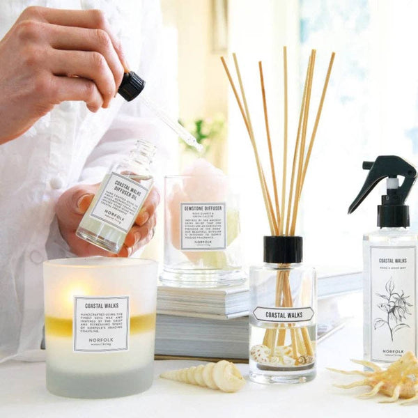 A person uses a dropper to add Norfolk Natural Living Coastal Walks Gemstone Diffuser Oil 50ml to a diffuser beside a lit candle, reed diffuser, spray bottle, and decorative seashells on a white table.