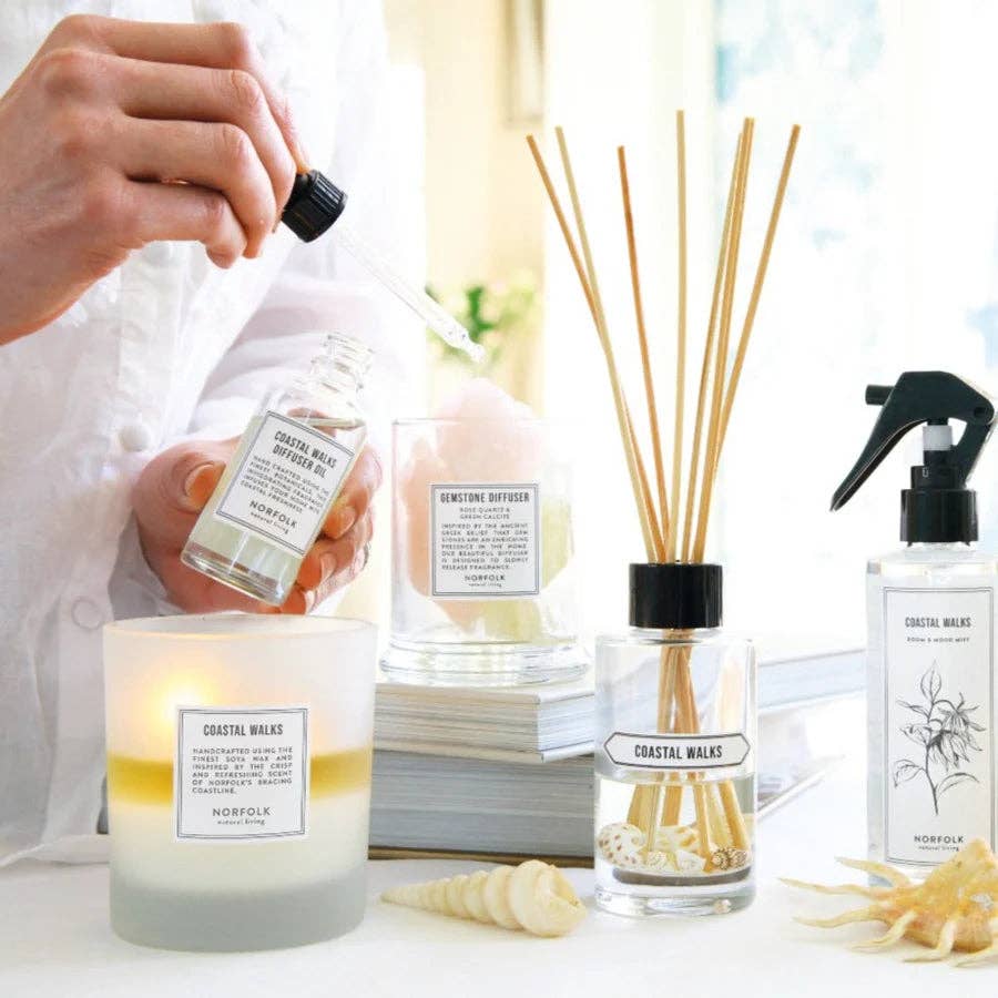 A hand holds a dropper over a bottle of Norfolk Natural Living Gemstone Diffuser Oil 50ml - Rose Garden, next to a candle, spray bottle, and seashell, all arranged on a white surface in bright natural light.
