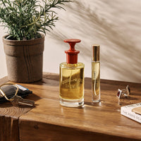 A Caswell-Massey Number Six Eau de Parfum 100mL bottle with a red cap rests on a wooden table beside a plant, keys, book, and cufflinks. Warm light highlights the classic fragrance by Caswell-Massey in this elegant scene.