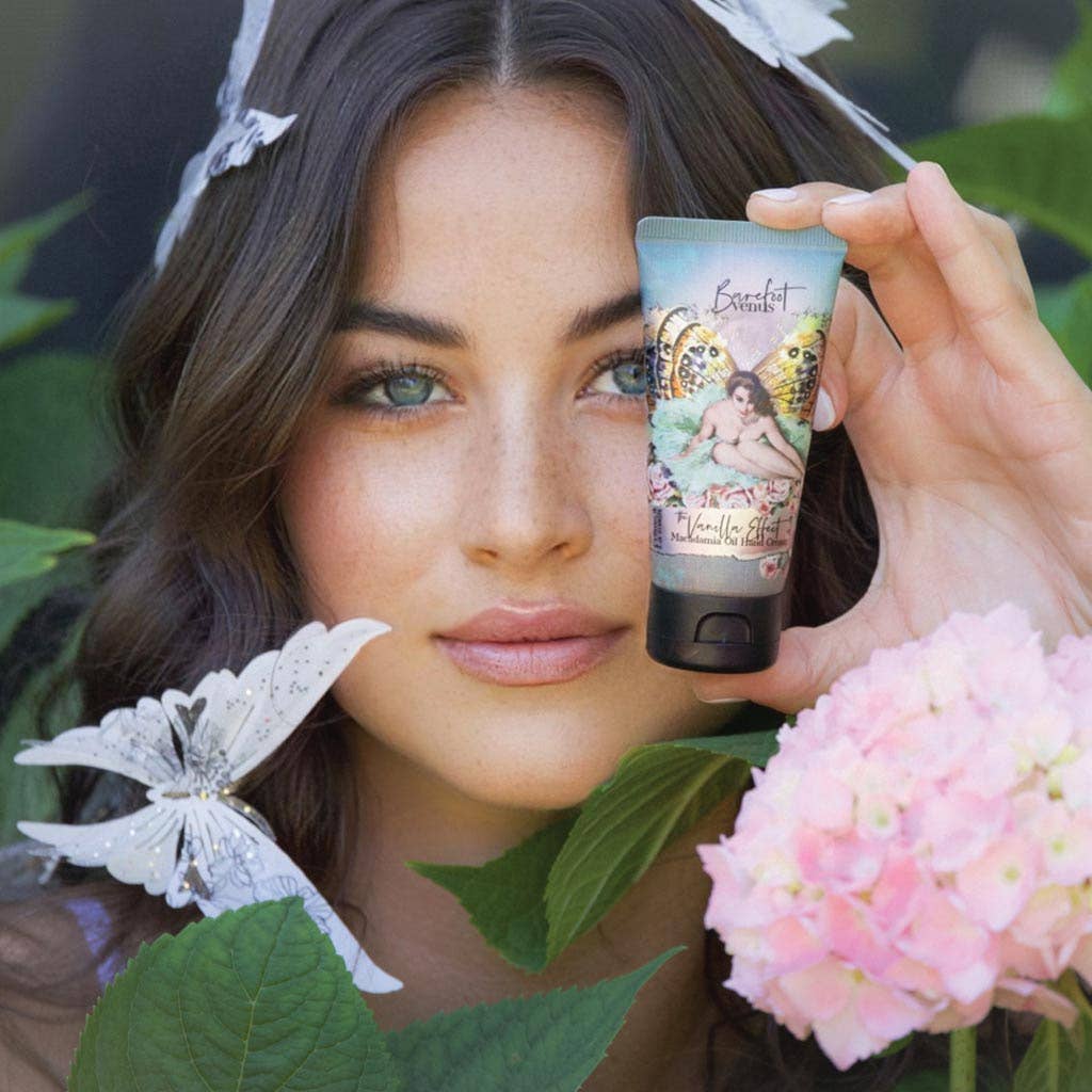 A person with long hair holds Barefoot Venus The Vanilla Effect Hand Cream, a cruelty-free beauty product, near their face. They are surrounded by green leaves and a pink hydrangea flower as a decorative white butterfly flits in the foreground.