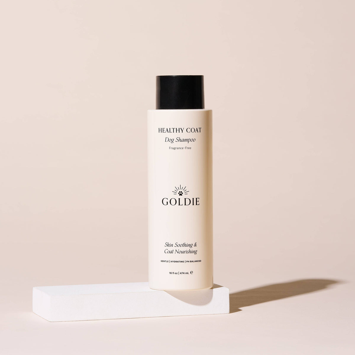 A bottle of Goldie - Healthy Coat by Goldie, featuring a black cap, stands upright on a white rectangular platform against a neutral beige background.