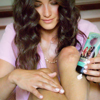 A woman with wavy brown hair applies Barefoot Venus Pink Pepper Lotion from a green tube onto her leg indoors. She wears a pink shirt, gold necklace, and has light pink painted nails.