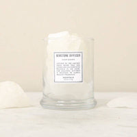 A clear glass container labeled "Norfolk Natural Living Gemstone Diffuser Snow Quartz" by Norfolk Natural Living holds large white aromatherapy stones on a light surface, surrounded by extra snow quartz pieces with a beige background.