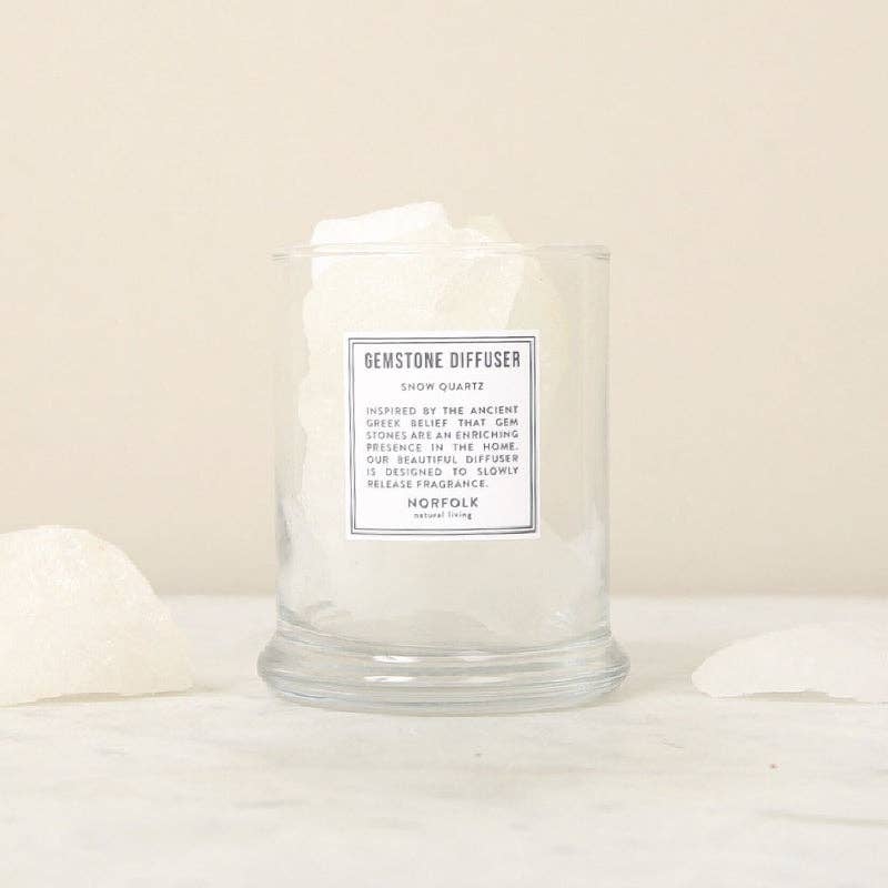 A clear glass container labeled "Norfolk Natural Living Gemstone Diffuser Snow Quartz" by Norfolk Natural Living holds large white aromatherapy stones on a light surface, surrounded by extra snow quartz pieces with a beige background.