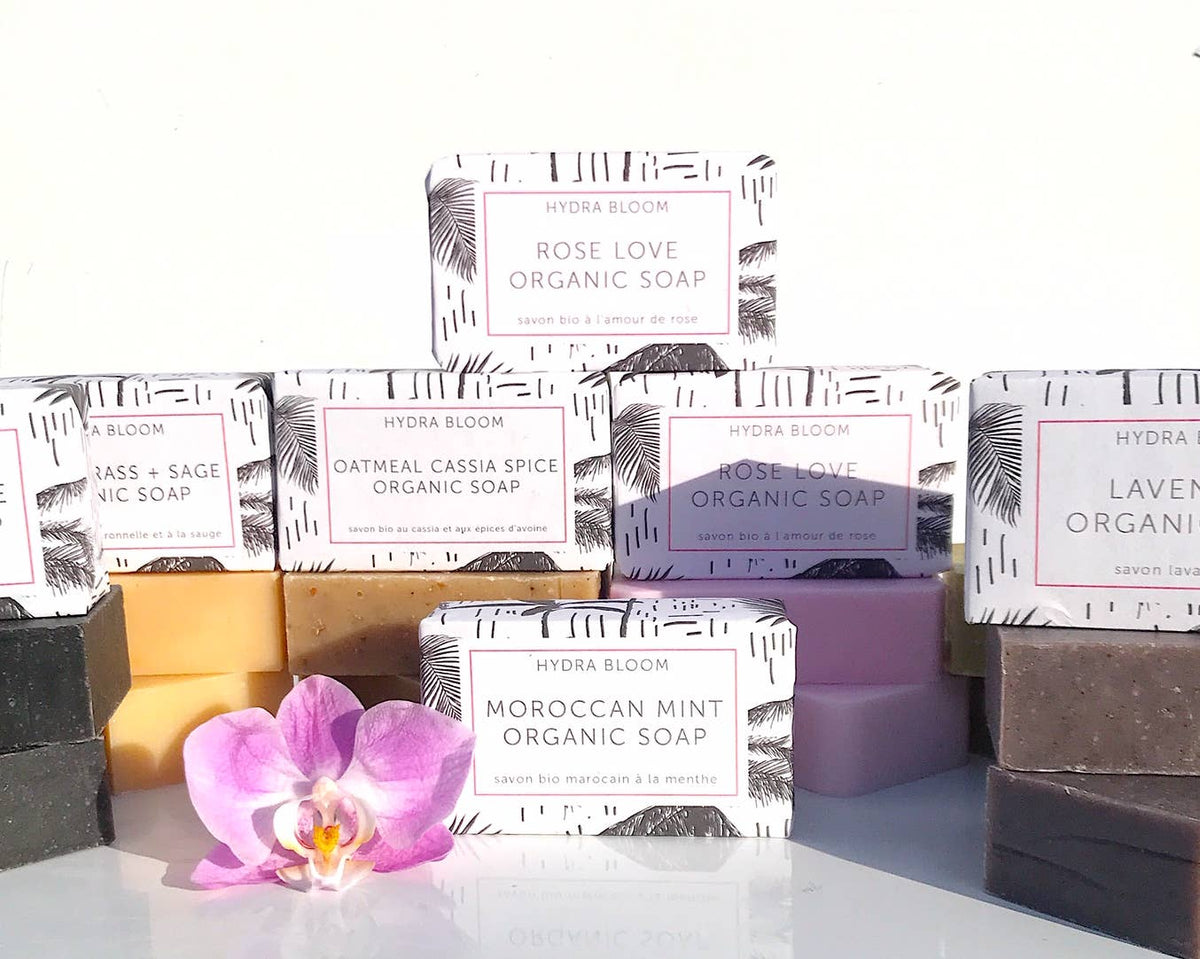 A collection of colorful Hydra Bloom Beauty Relax Lavender Organic Soap bars displayed with their packaging, featuring various scents such as lavender love and Moroccan spice, highlighted by an orchid flower in front.