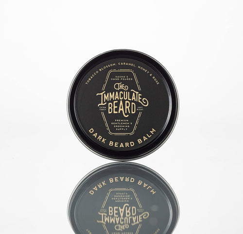 A round tin of The Immaculate Beard DARK Beard Balm by The Immaculate Beard rests on a white reflective surface. Its handmade label features a tobacco caramel scent with honey and rose notes, highlighting its hand-crafted quality.