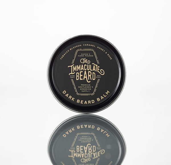 A round tin of The Immaculate Beard DARK Beard Balm by The Immaculate Beard rests on a white reflective surface. Its handmade label features a tobacco caramel scent with honey and rose notes, highlighting its hand-crafted quality.