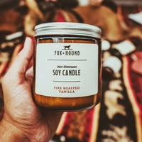 A hand holding a "Fox + Hound Odor Eliminator Soy Candle - Fire Roasted Vanilla" with a "Fire Roasted Vanilla" label, in front of a blurred colorful background.