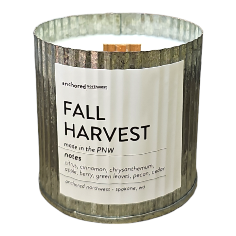 Hand-poured in a metal container, the Anchored Northwest Fall Harvest Rustic Vintage Farmhouse Wood Wick Candle features a wooden wick. The label reads "anchored northwest FALL HARVEST made in the PNW." Scent notes include citrus, cinnamon, chrysanthemum, apple, berry, green leaves, pecan, and cedar. Produced by Anchored Northwest in Spokane, WA with 60 hours of burn time.