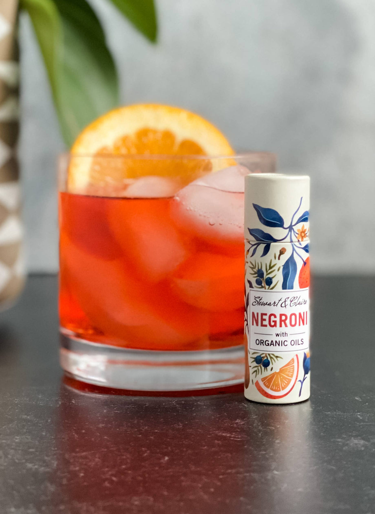 A glass of Negroni with ice and orange sits beside a Stewart & Claire Negroni Lip Balm in a paper tube, labeled "eco-friendly lip care," on a dark surface with a leafy plant in the background.