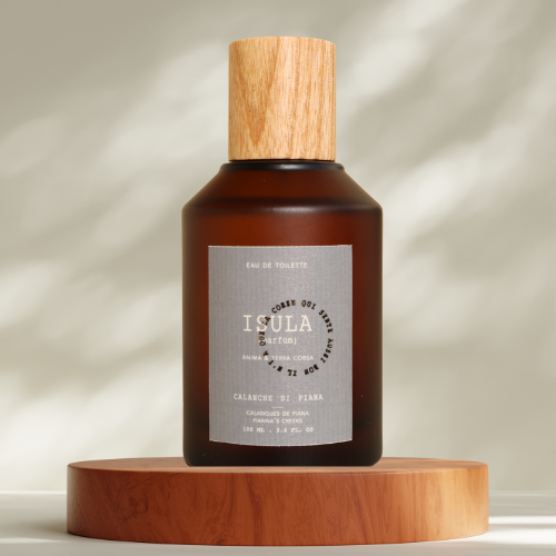 A brown glass ISULA parfums Calanque de Piana eau de toilette bottle with a wooden cap rests on a round wooden platform. The label reads "ISULA parfums," evoking Mediterranean notes and nature-inspired light patterns in the background.