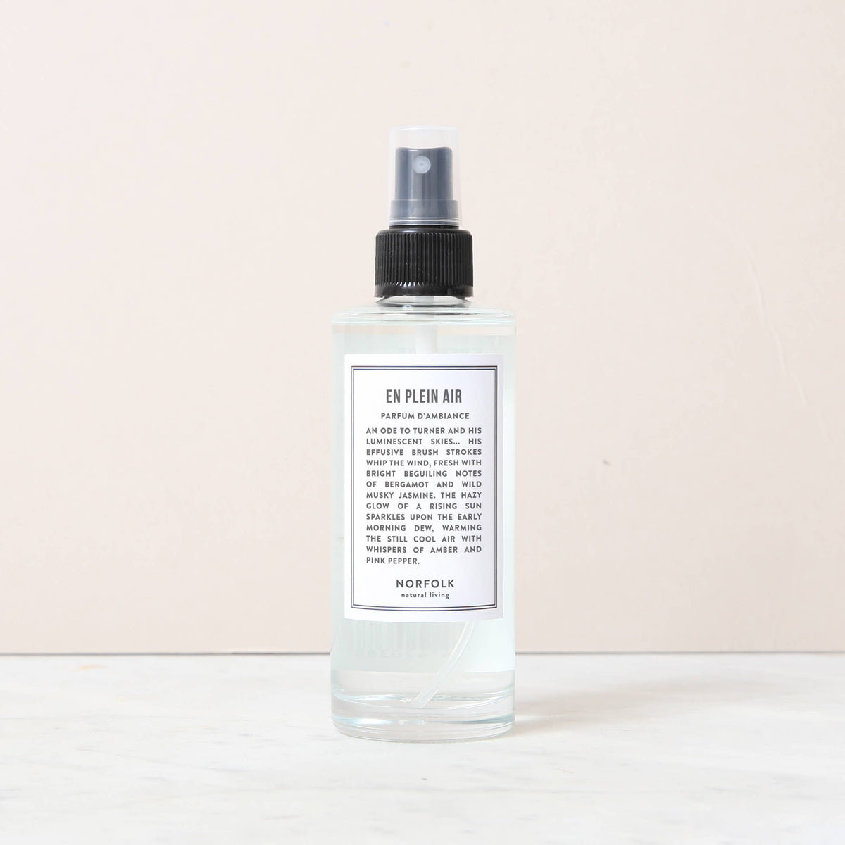 A clear 150ml Norfolk Natural Living En Plein Air Room and Mood Mist, featuring a transparent liquid and sleek black nozzle, rests on white marble with a light beige background.