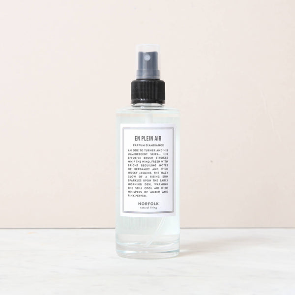 A clear 150ml Norfolk Natural Living En Plein Air Room and Mood Mist, featuring a transparent liquid and sleek black nozzle, rests on white marble with a light beige background.