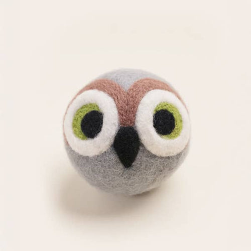 The MODERNBEAST - OWL BEASTBALL Wool Dog Toy is a handmade, round felted wool ball with large green and white eyes, a black beak, and brown markings above the eyes on a gray background.