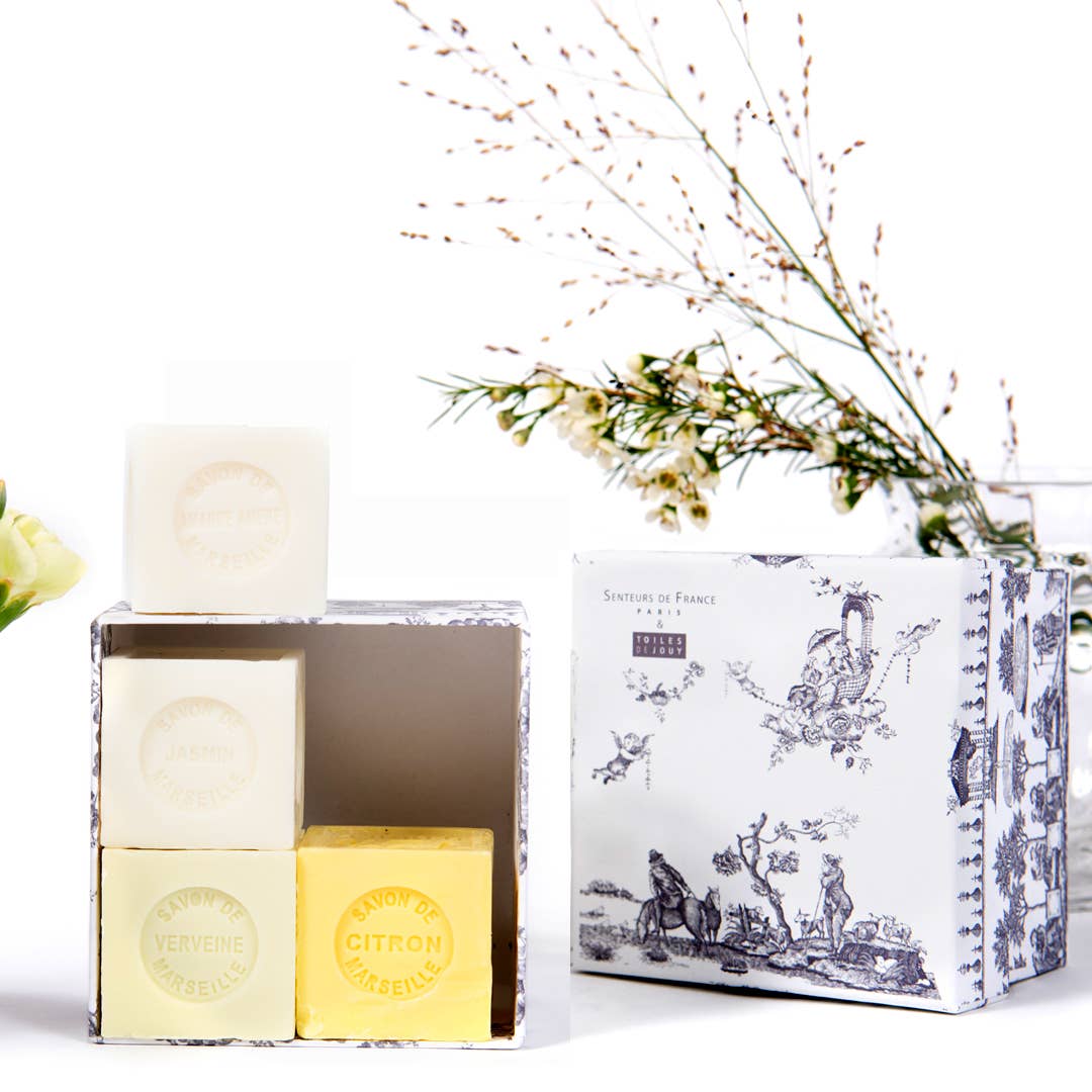 Three pastel Senteurs De France cube soaps—Honeysuckle, Almond, Jasmine, and Verbena—are elegantly stacked in a decorative French box, with floral accents and greenery highlighting this artisanal soap collection.