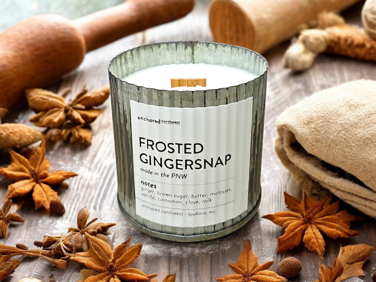 An "Anchored Northwest Frosted Gingersnap Rustic Vintage Farmhouse Wood Wick Candle" by Anchored Northwest sits on a wooden surface surrounded by star anise, cinnamon sticks, and fabric rolls. The candle's label mentions scents of ginger, brown sugar, butter, molasses, vanilla, cinnamon, clove, and milk.