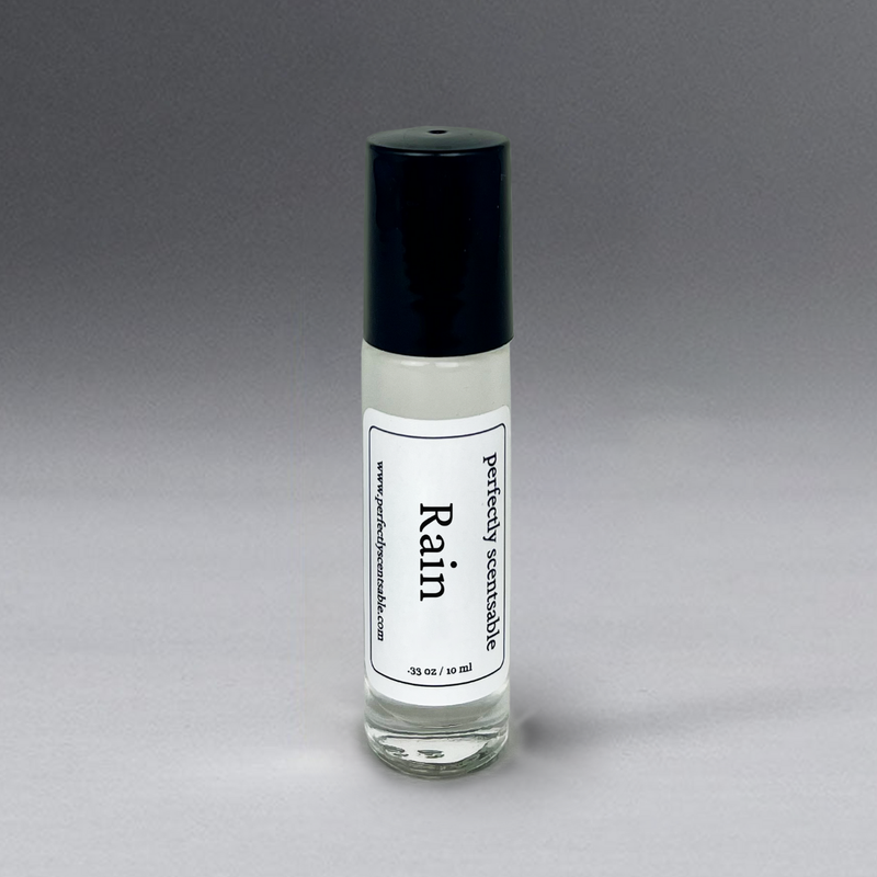 A small glass bottle with a black cap is filled with a clear liquid. The label on the bottle reads "Perfectly Scentsable Rain Perfume Oil - Rollerball" and indicates it contains 0.35 oz / 10 ml of Rain Perfume Oil, a gender-neutral scent from Perfectly Scentsable. The background is a gradient of gray, transitioning from light at the bottom to darker at the top.