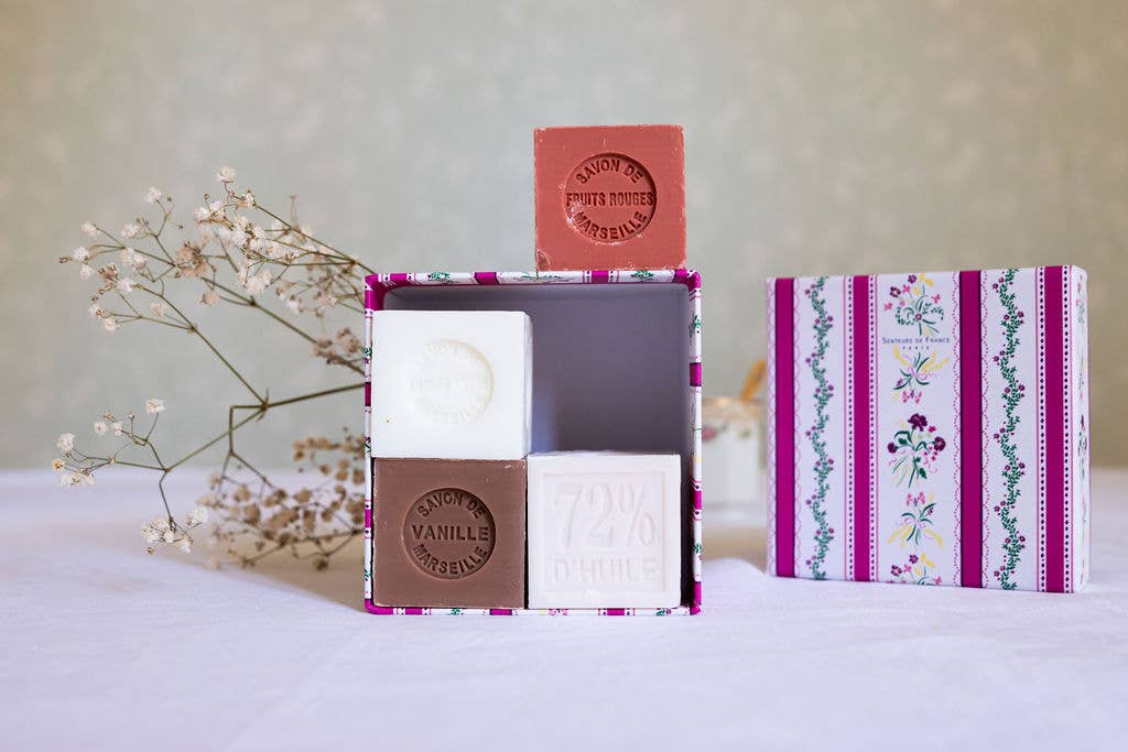 The Senteurs De France Coffret features four artisanal Marseille cube soaps—Almond, Rose, Fig, and Vanilla—in a decorative floral-striped box by Senteurs De France, with dried white flowers adding charm on a white tablecloth.