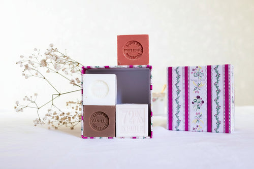 The Senteurs De France Coffret features four artisanal Marseille cube soaps—Almond, Rose, Fig, and Vanilla—in a decorative floral-striped box by Senteurs De France, with dried white flowers adding charm on a white tablecloth.
