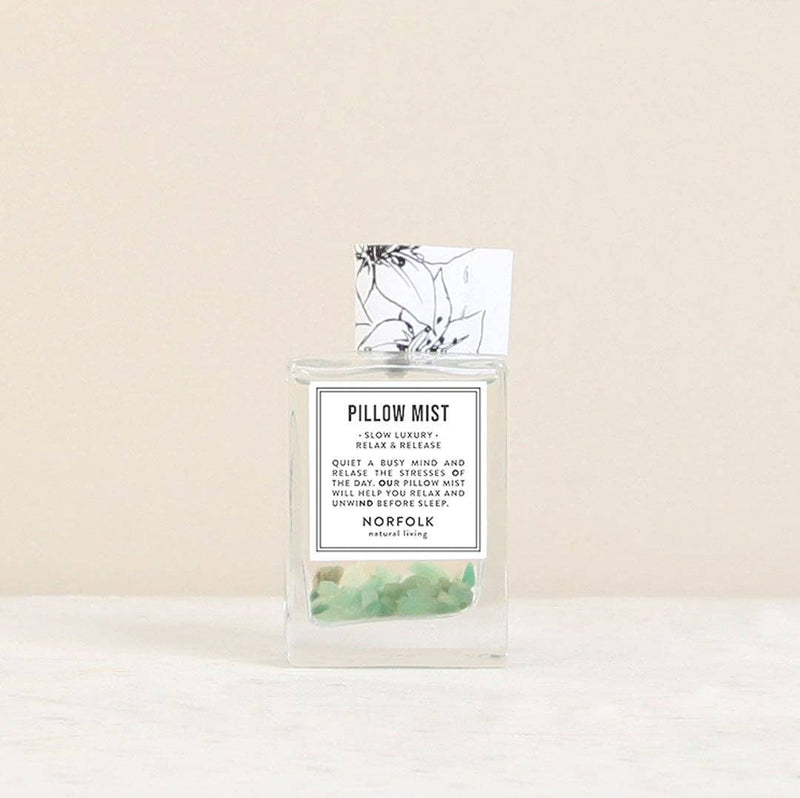 The Norfolk Natural Living Relax and Release Pillow Mist (50ml) is a clear glass bottle with essential oils and green stones, featuring a black-and-white floral card behind the label, all set against a light backdrop.