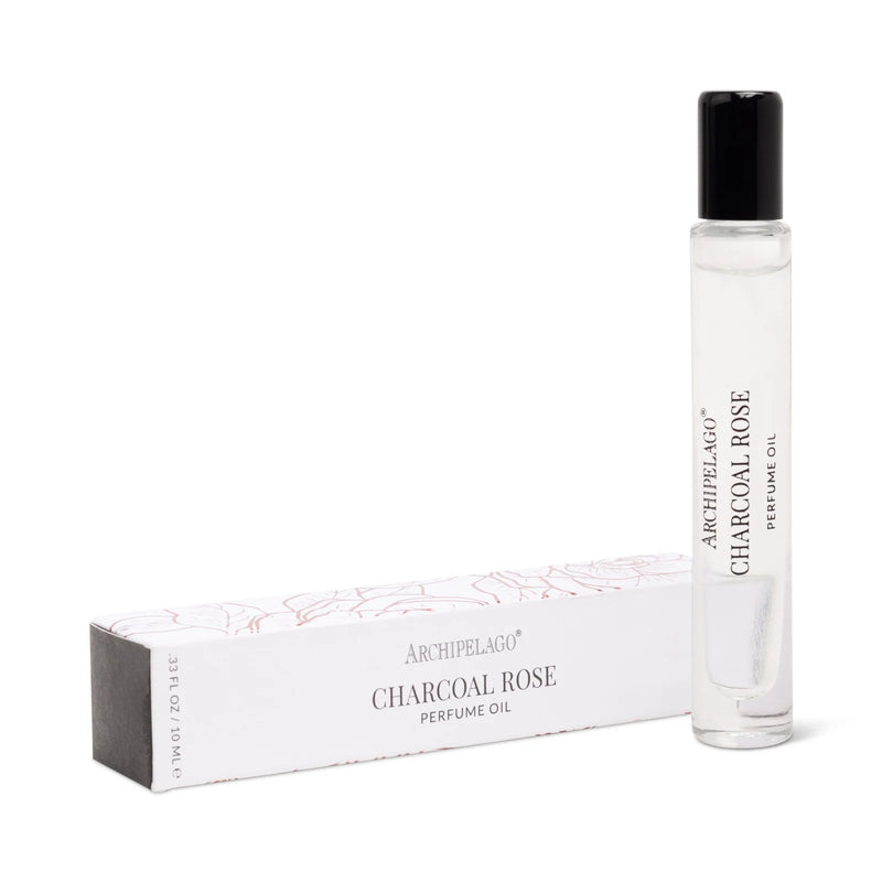 A clear glass rollerball bottle of Archipelago Botanicals Charcoal Rose Perfume Oil stands upright beside its white box with pink accents—a chic choice for fans of long-lasting fragrance.
