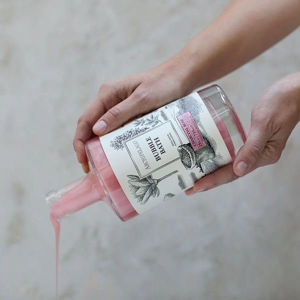 A person pours thick pink liquid from an Archipelago Botanicals Charcoal Rose 25 oz. Luxury Bubble Bath glass bottle, adorned with floral illustrations, against a neutral textured background.
