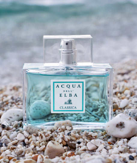 A bottle of Acqua dell'Elba Classica Uomo Eau de Parfum - 100ml by Acqua dell'Elba sits on white pebbles at the beach, evoking Mediterranean fragrance as the sea blurs gently in the background.